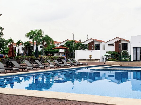 A swimming pool with lounge chairs and umbrellas, surrounded by buildings and greenery.