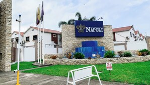 Señalización para 'Urbanización Napoli', con un muro de piedra, una valla blanca y un área con césped.