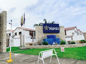 A sign for 'Urbanización Napoli' with a stone wall, a white fence, and a grassy area.