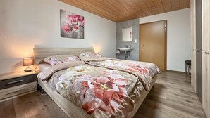 Une chambre avec un plafond en bois, un lit recouvert d’une literie à motifs floraux, une table de chevet avec une lampe, un miroir fixé au mur et une salle de bain avec un lavabo et des toilettes.