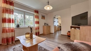 Un salon chaleureux avec une table basse en bois, un canapé et un téléviseur. On aperçoit une fenêtre avec des rideaux, un radiateur et, au fond, un coin cuisine.