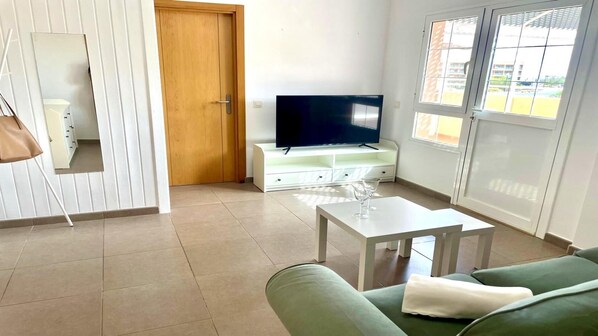 Una sala de estar con televisor sobre un mueble blanco, un sofá verde y una mesita con vasos.