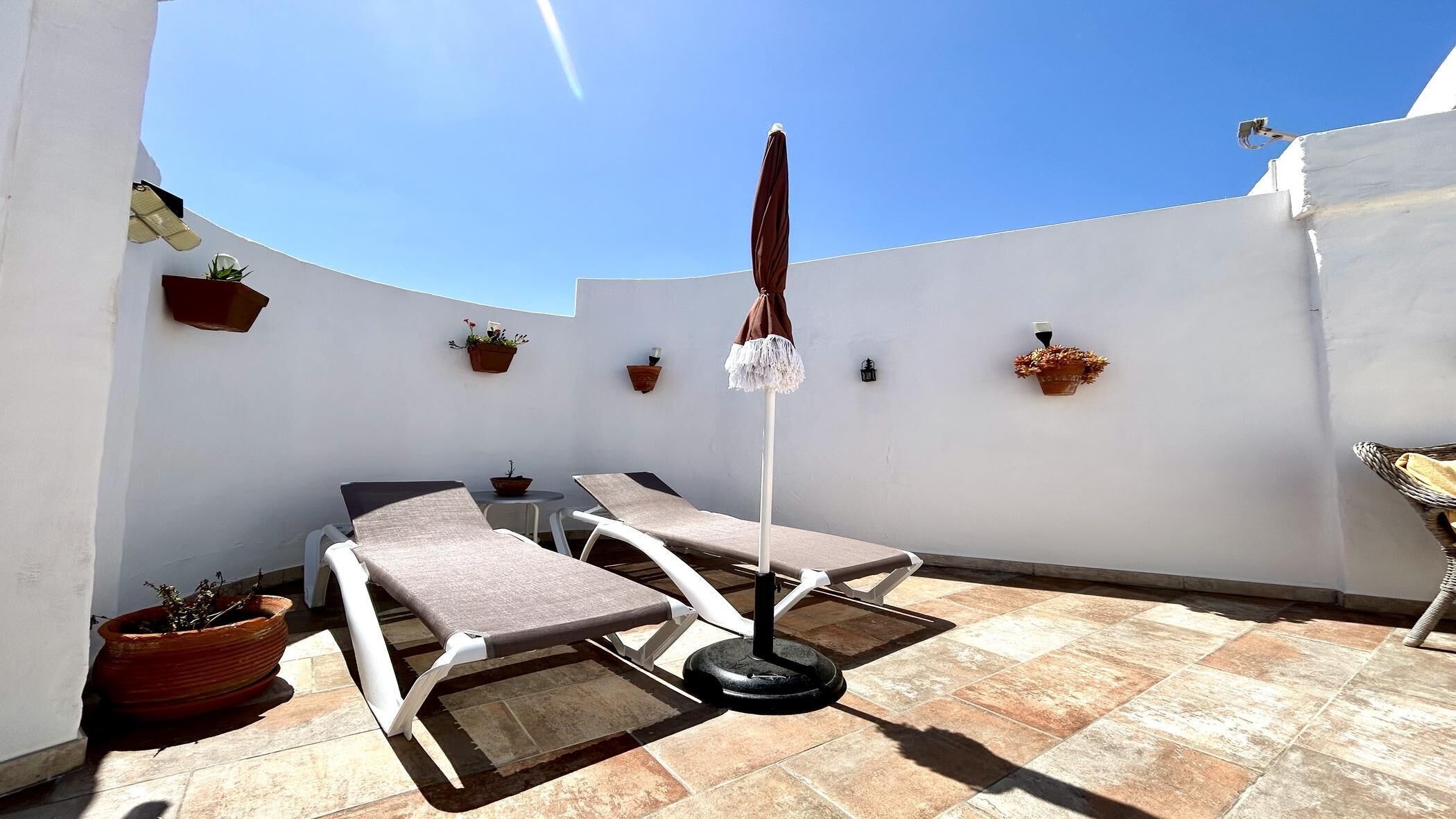 Een zonnig buitenterrein met loungestoelen en één parasol, omringd door witte muren en potplanten.