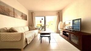 Una sala de estar con un sofá, una mesa de centro, un televisor sobre un mueble de madera y vistas al exterior.