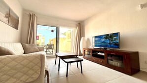 Una sala de estar con un sofá, una mesa de centro, un televisor sobre un soporte de madera y vista a un edificio amarillo a través de una puerta de vidrio.