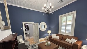 A living room with a brown sofa, a white armchair, a coffee table, a chandelier, and a large window with blinds. - "GraciaDeDios" Beautifully renovated 1910 Cottage (Mount Barker)