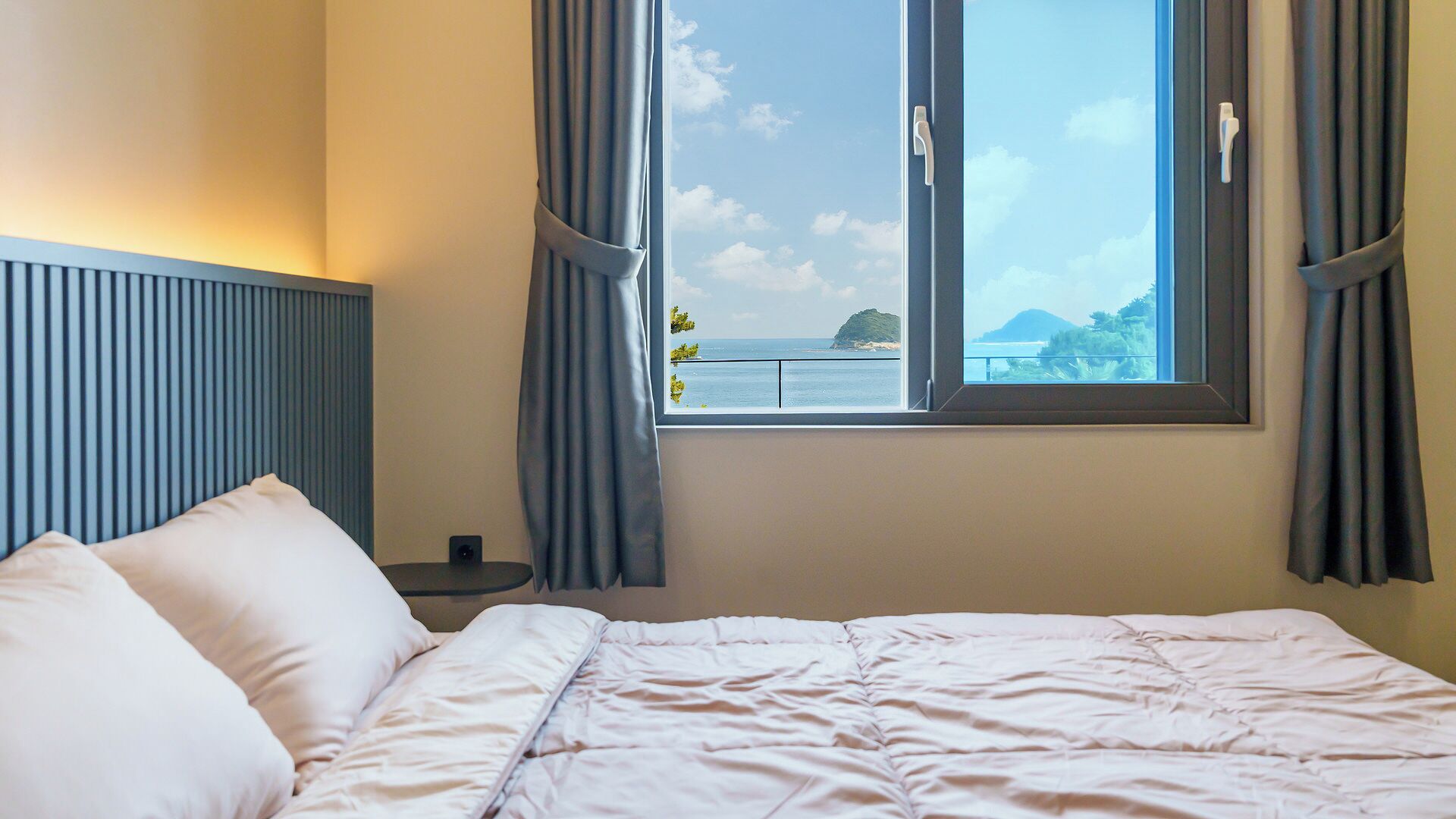 A hotel room with a bed, a nightstand, a window with curtains, and a view of the sea.