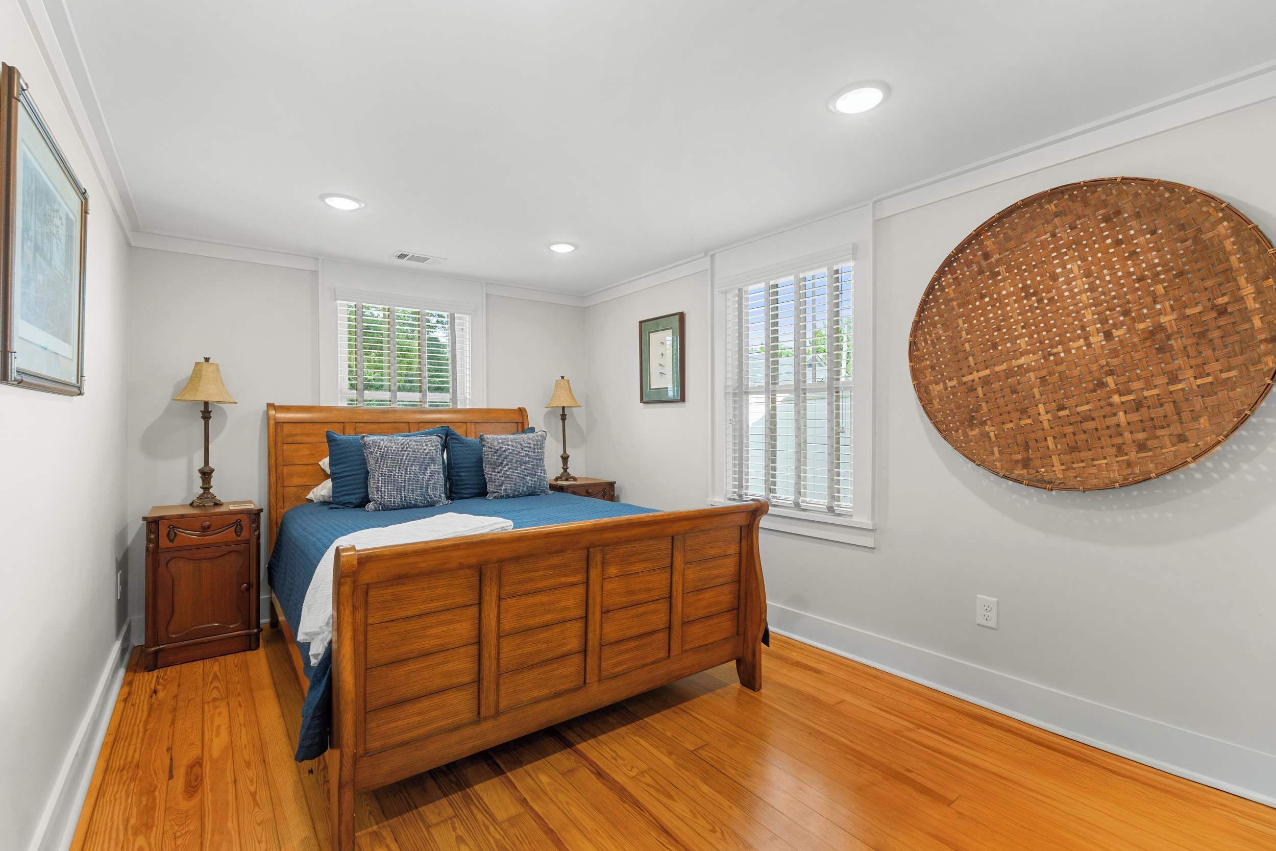A bedroom with a wooden bed, two bedside tables, a round woven wall hanging, and windows with blinds.