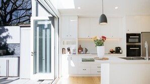 Een moderne keuken met witte kasten, een groot eiland, een hangende lamp en uitzicht op de buitenlucht.