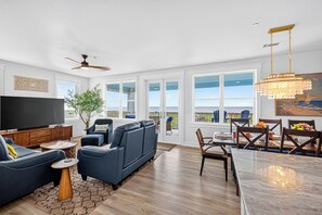 Een ruime woonkamer met een grote flatscreen-tv, een plafondventilator en een eethoek met uitzicht op de oceaan.