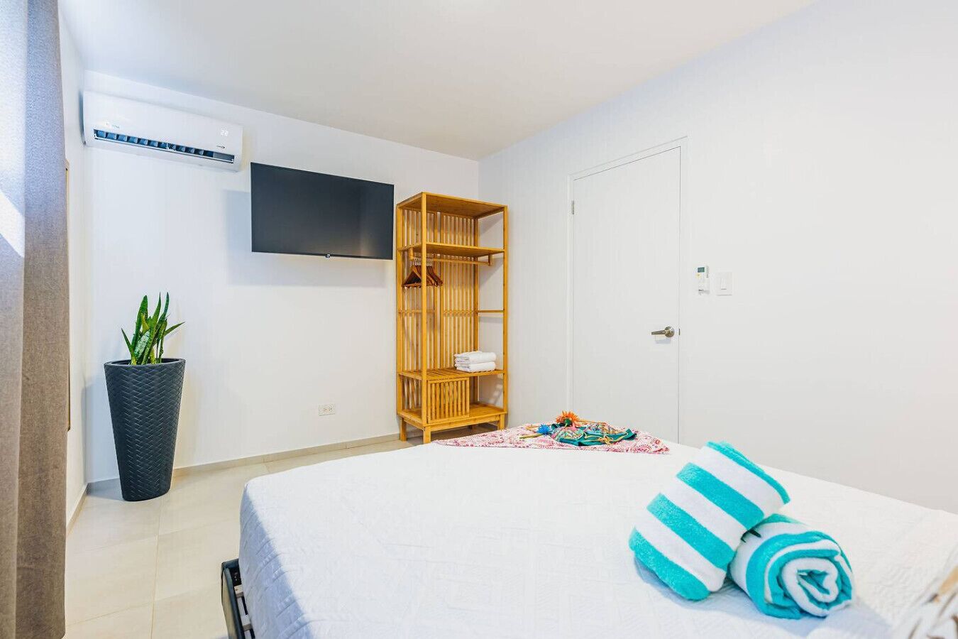 Un dormitorio con una cama, un televisor en la pared, un armario de madera, una planta en maceta y una puerta.