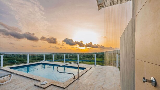 Une piscine sur le toit offrant une vue sur le coucher de soleil sur l’océan, entourée d’un bâtiment moderne doté d’un sol carrelé et d’une rambarde en métal.