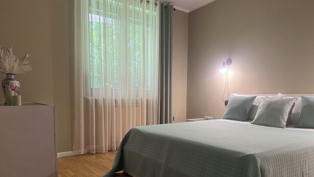 Ein Schlafzimmer mit einem Bett, einem Nachttisch, einer Lampe und einem Fenster mit durchsichtigen Vorhängen.