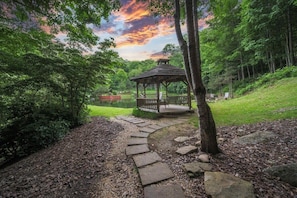 Un kiosque dans un parc, accessible par un chemin de pierre, entouré d’arbres et bordé d’un lac.