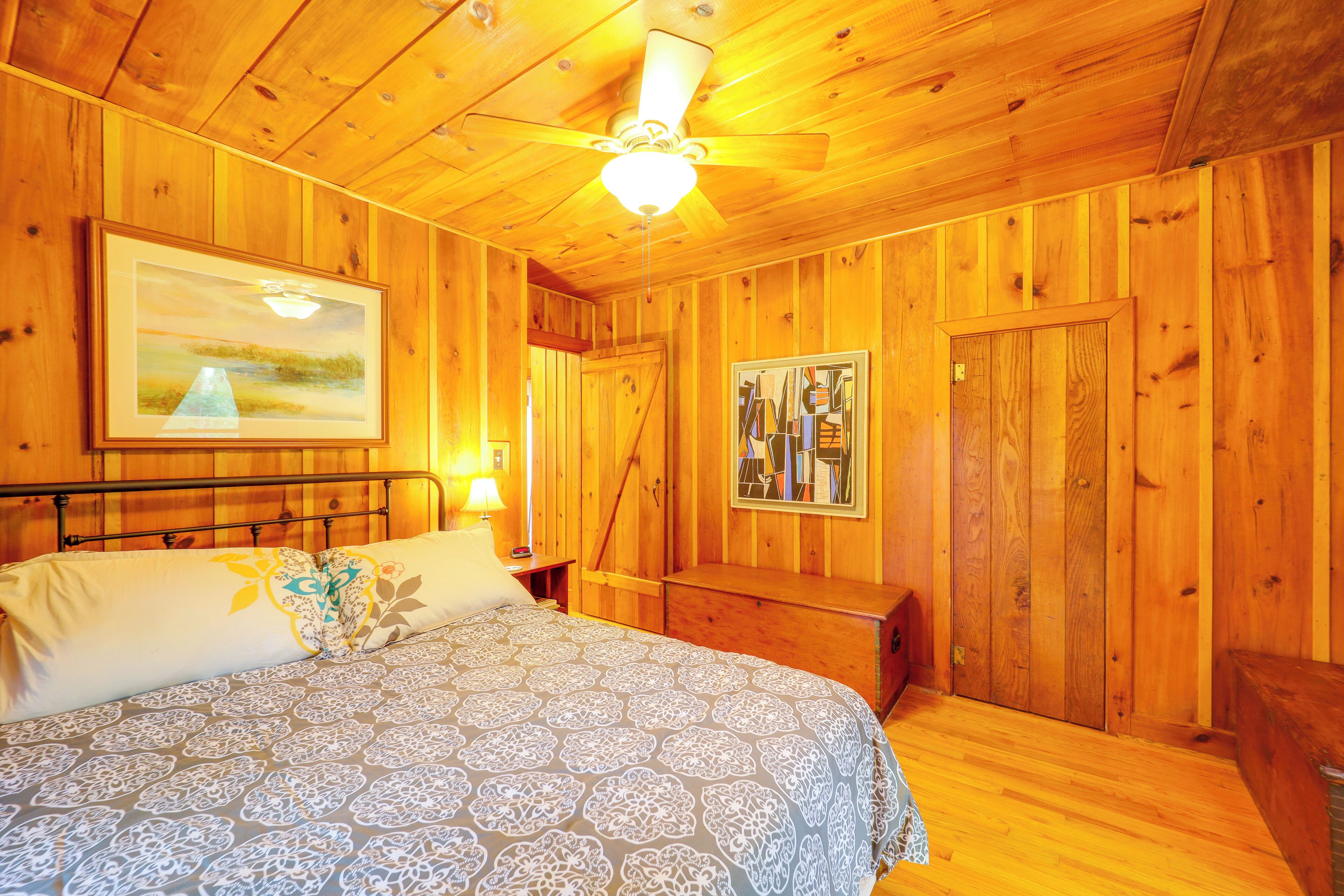 Une chambre à coucher dans une cabane en bois, avec un lit, une table de chevet, un ventilateur de plafond et un tableau accroché au mur.
