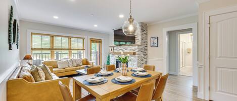 A dining area with a wooden table set for four, surrounded by chairs. There is a stone fireplace with a chimney, a hanging light fixture, and a view of the outdoors through large windows.