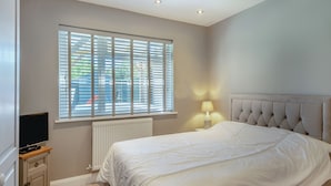 Een slaapkamer met een bed, een nachtkastje met een lamp, een televisie op een meubelstuk en een raam met jaloezieën.
