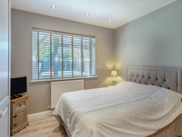 A bedroom with a bed, a nightstand with a lamp, a TV on a stand, and a window with blinds. - 2 bedroom accommodation in Snettisham (Snettisham)