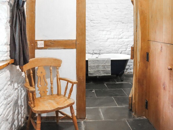 A narrow hallway with a wooden chair, a white wall, a wooden door, and a bathroom with a bathtub and a sink.