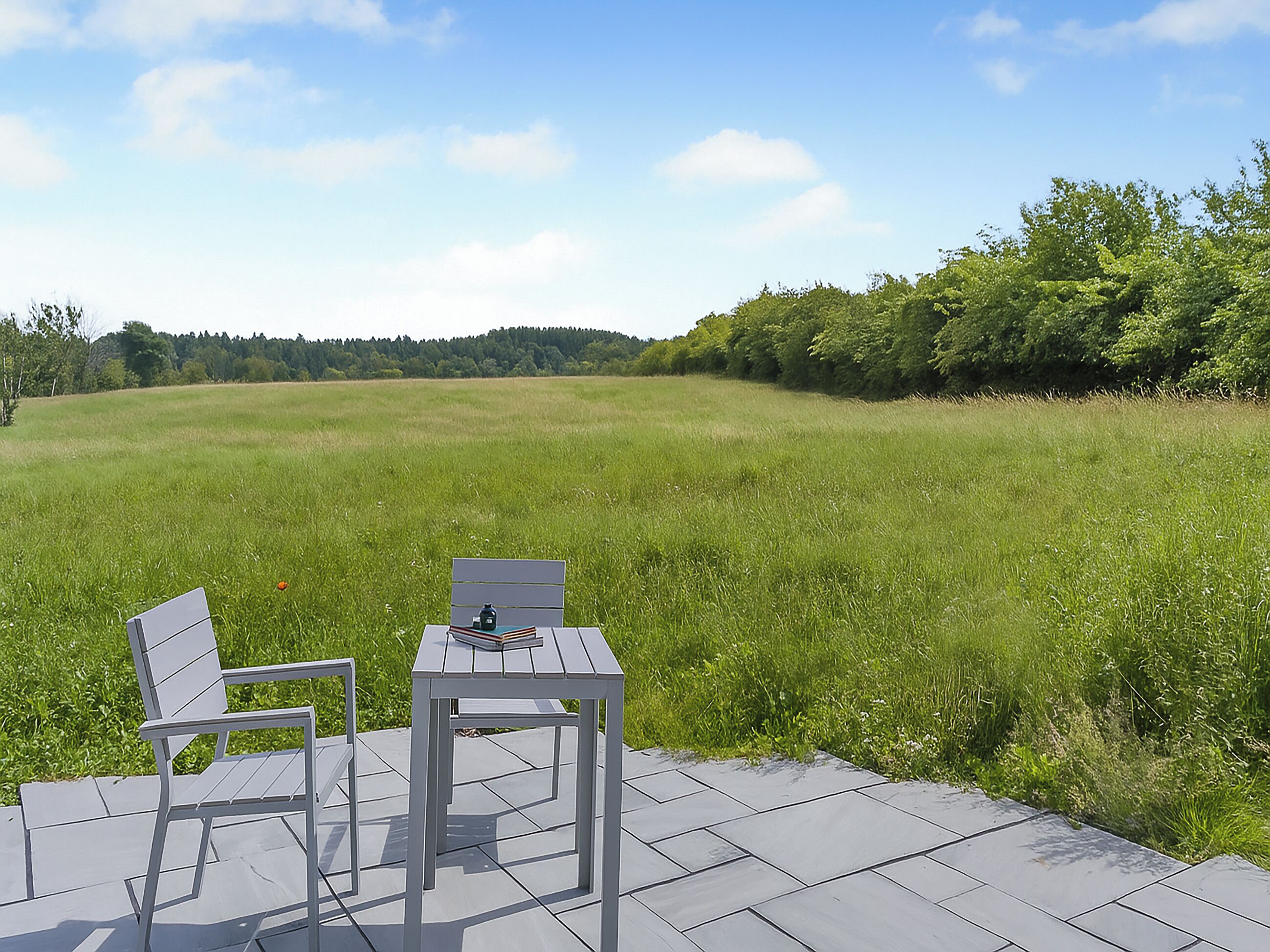 An outdoor seating area with a table and two chairs on a tiled patio overlooking a grassy field.