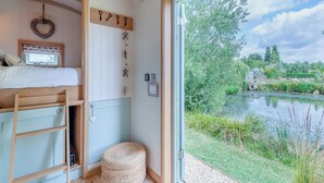 A bedroom with a bunk bed, a small round ottoman, a wall-mounted shelf, and a window with a view of a lake and greenery. - Herbie's Shepherd Hut - UK50229 (Nottingham)