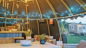 A cozy tent with a wooden interior, string lights, a bar area, and a view of a wooden house outside. - Herbie's Shepherd Hut - UK50229 (Nottingham)