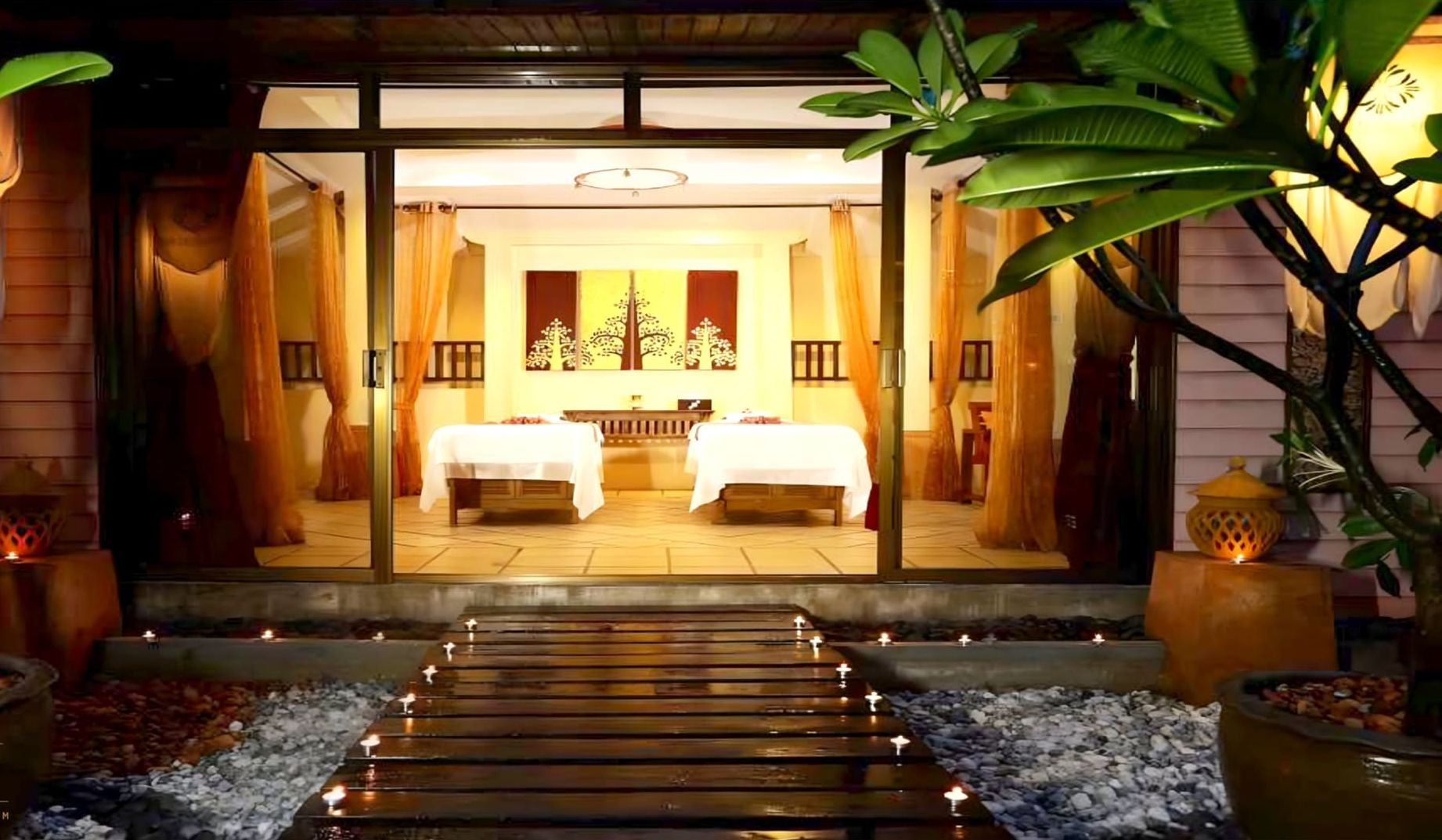 A hotel entrance with a pathway leading to a room with two beds, a reception area, and tropical plants.
