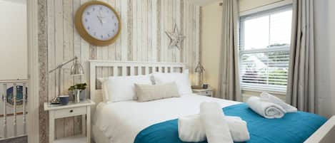 Un lit bien fait, avec une couvre-lit bleue et des oreillers blancs, une horloge ronde accrochée au mur, une décoration en forme d’étoile et une fenêtre avec des rideaux.