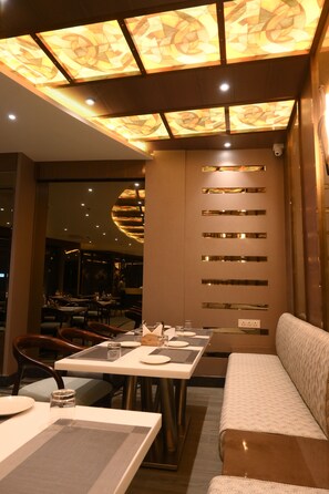 A restaurant interior with a modern design, featuring a patterned ceiling, a seating area with tables and chairs, and a wall with horizontal metallic strips. - Amara Super Deluxe Dormitory (Varanasi)