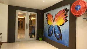Un vestíbulo moderno con entrada por puerta de cristal, un mural grande de mariposas, una planta en maceta y un objeto circular rojo en la pared.