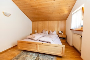 A bedroom with a wooden ceiling, a bed with floral bedding, a nightstand, a bedside table, a window with curtains, and a radiator.
