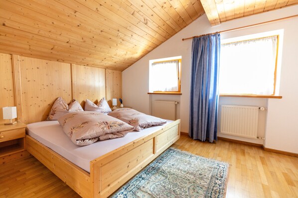 Een houten kamer met een bed, nachtkastje, raam met gordijnen en een radiator.