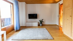 A room with a wooden ceiling, a flat-screen TV mounted on the wall, a white cabinet, a potted plant, and a large rug on the floor.