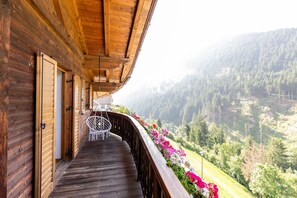 Un balcon en bois, avec une chaise blanche et une table, donnant sur une vallée boisée.