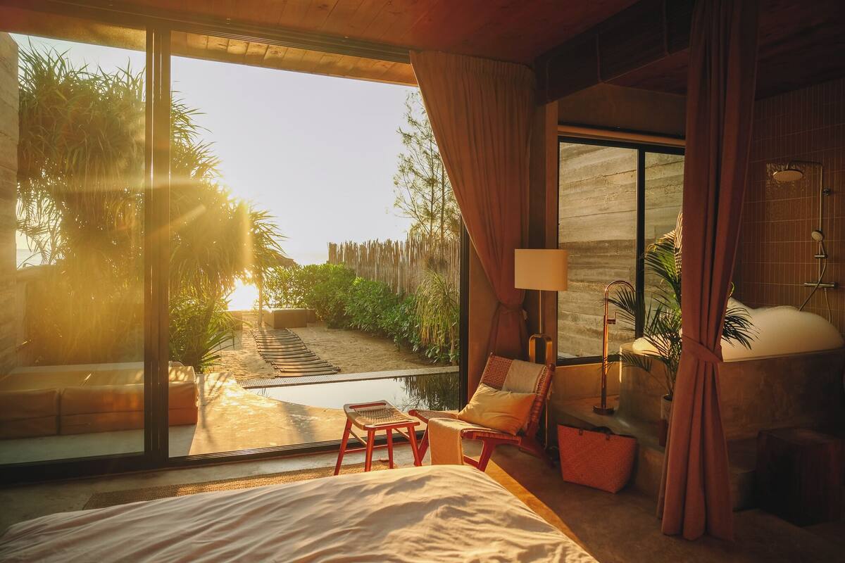 A bedroom with a large sliding glass door leading to a patio, a bed, a chair, a small table, and a bathtub.