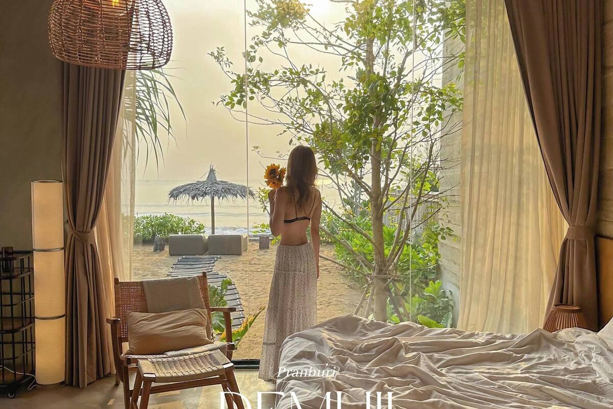 A bedroom with a view of a beach and a tree. There is a bed, a chair, and a hanging light fixture.
