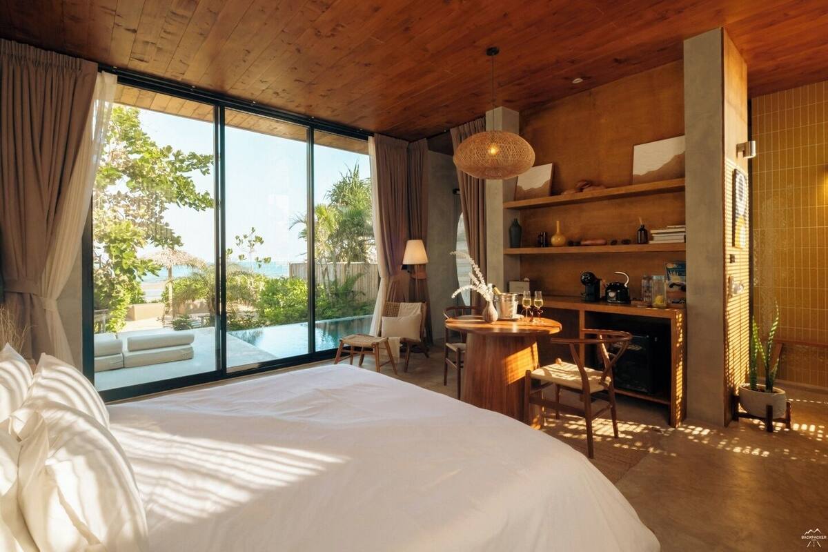 A modern hotel room with a large bed, a dining area, and a view of the outdoors.