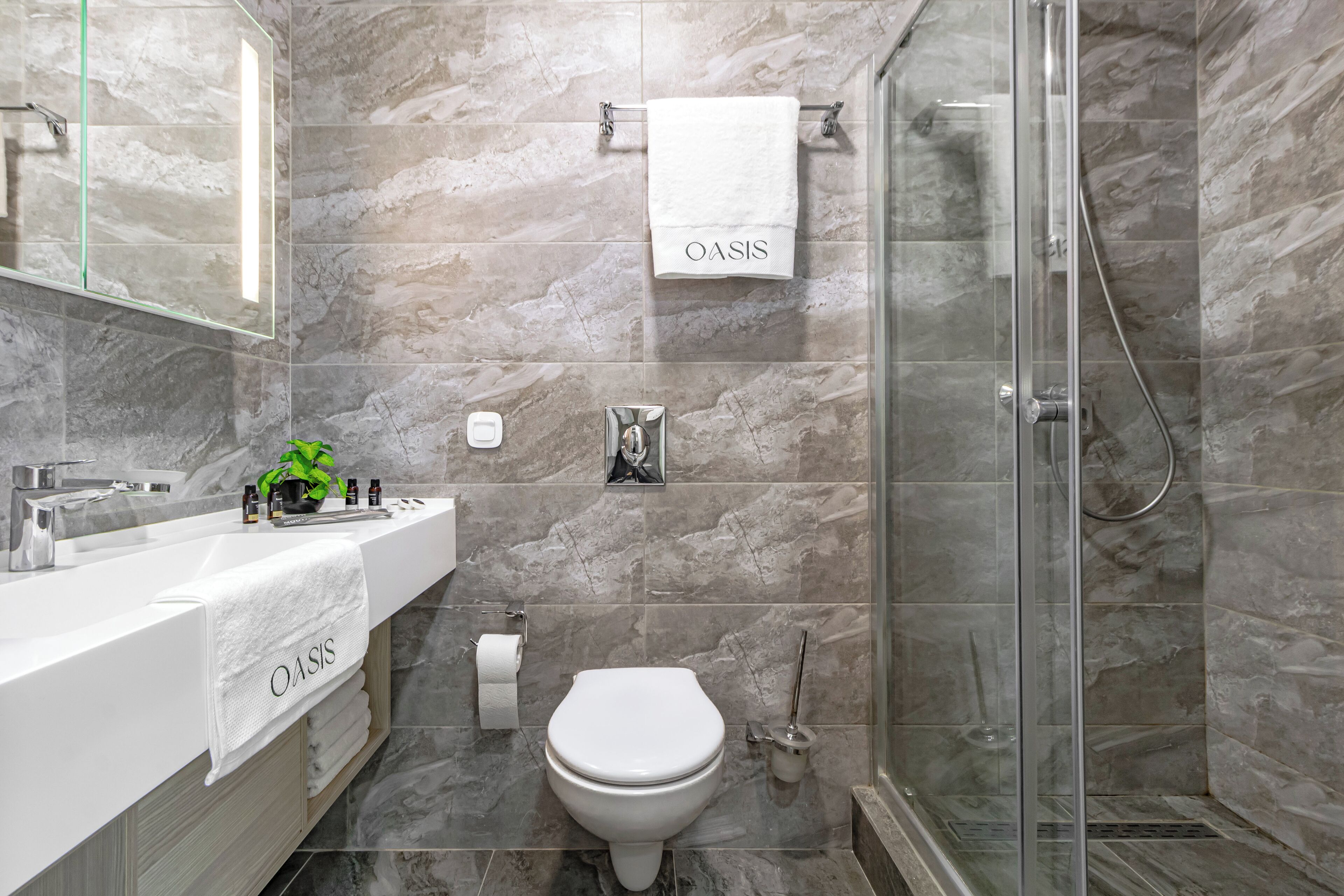 A modern bathroom with a marble backsplash, a glass shower enclosure, a white sink with a towel labeled "OASIS," and a toilet.