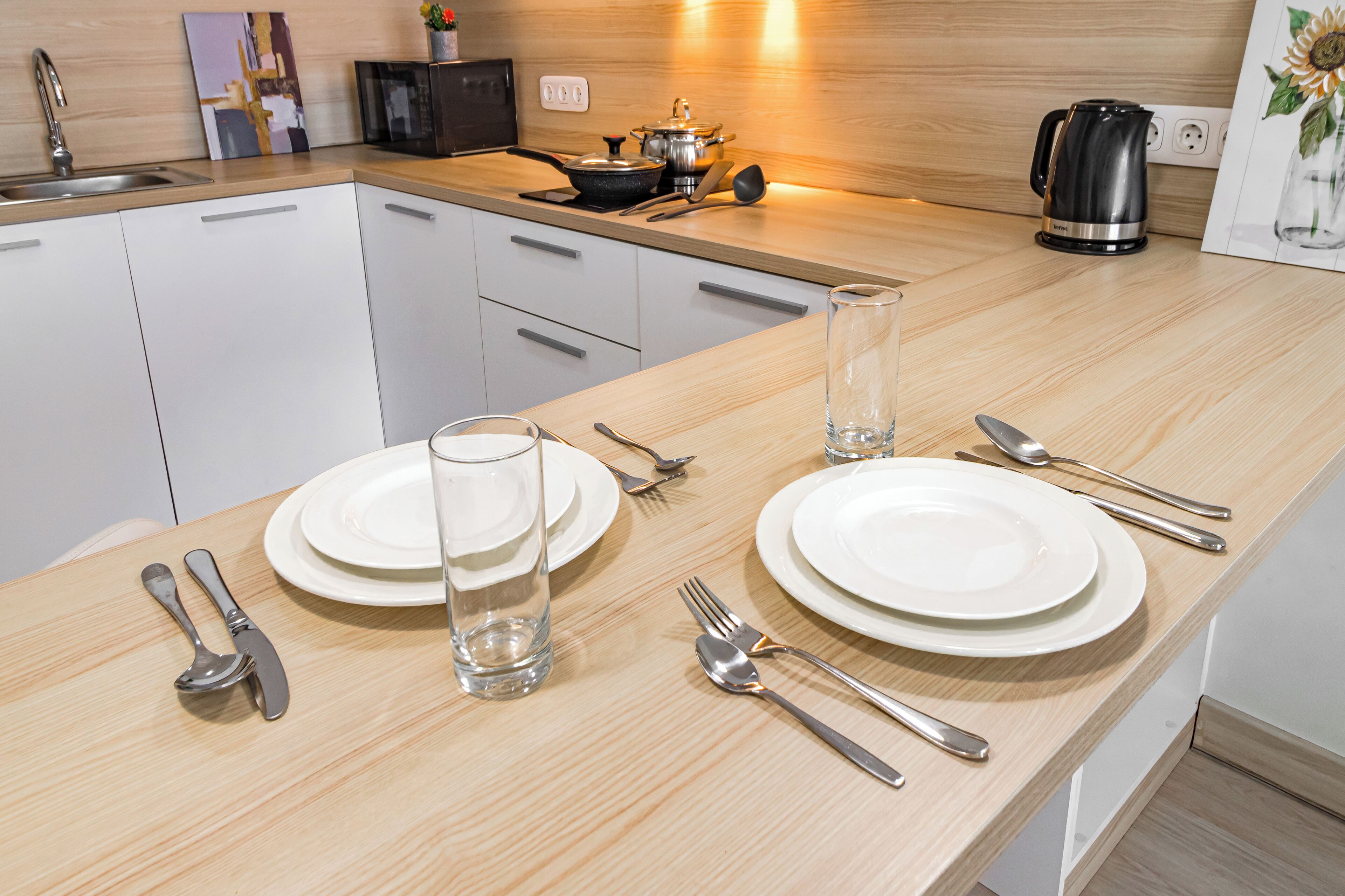 A modern kitchen with white cabinets, a wooden countertop, and a dining area set with white plates, cutlery, and a glass.