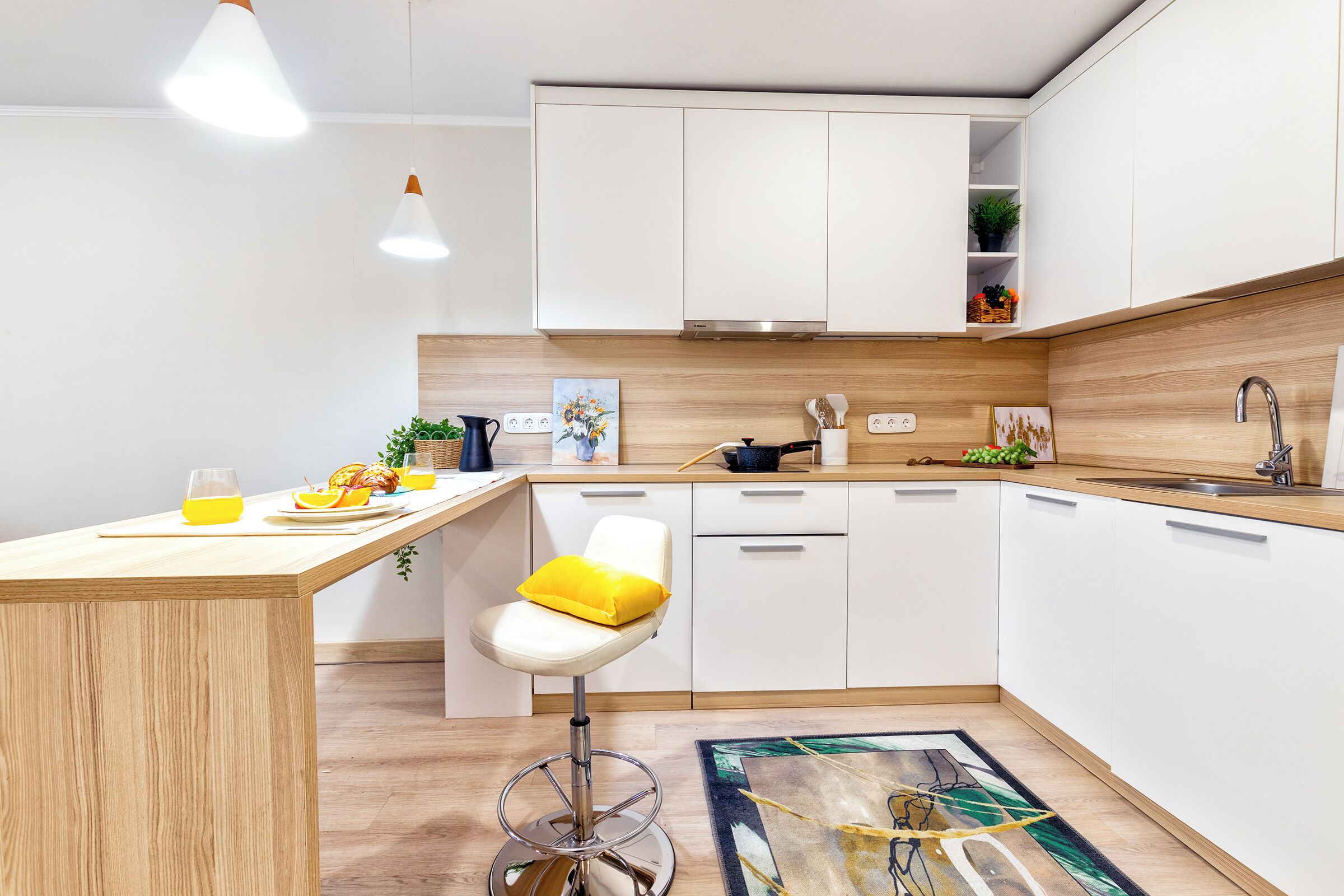 Una cucina moderna con mobili bianchi, un piano di lavoro in legno, uno sgabello da bar con cuscino giallo e un tappeto colorato.