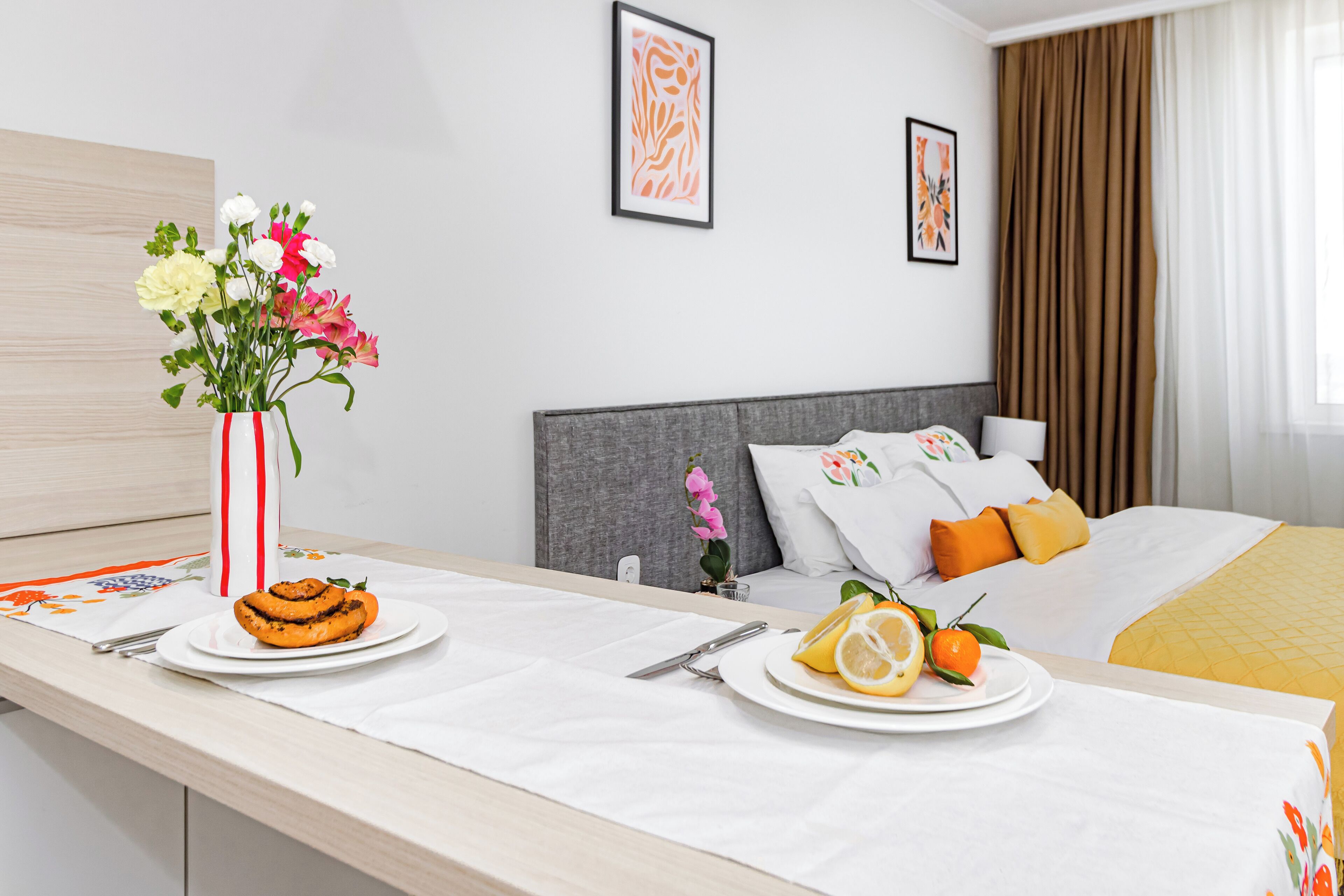 A hotel room with a bed, a bedside table with breakfast items, and a vase of flowers.