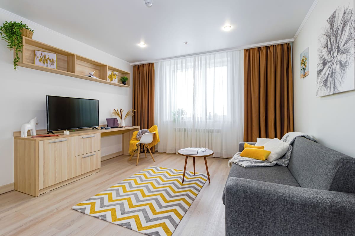A modern living room with a flat-screen TV, a grey sofa, a wooden desk, and a dining area with a yellow chair.