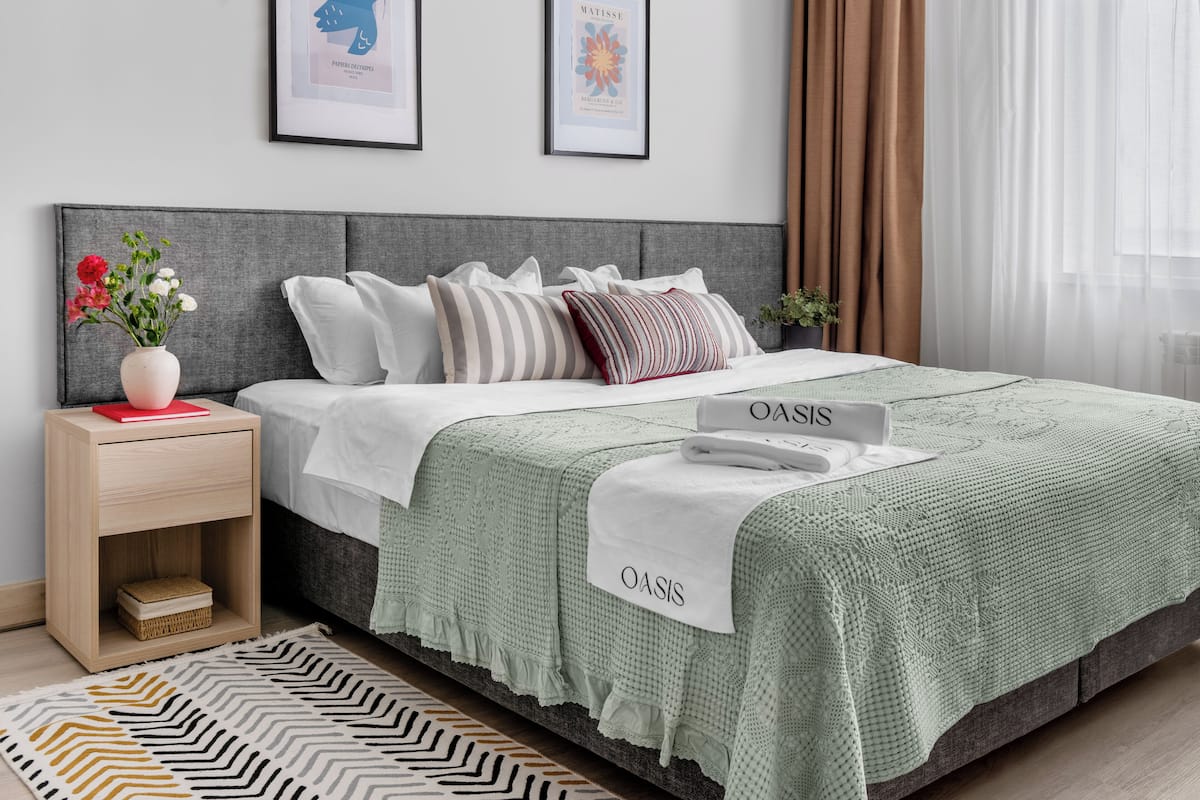 A neatly arranged bedroom with a grey headboard, a green textured bedspread, and a bedside table with a vase of flowers.