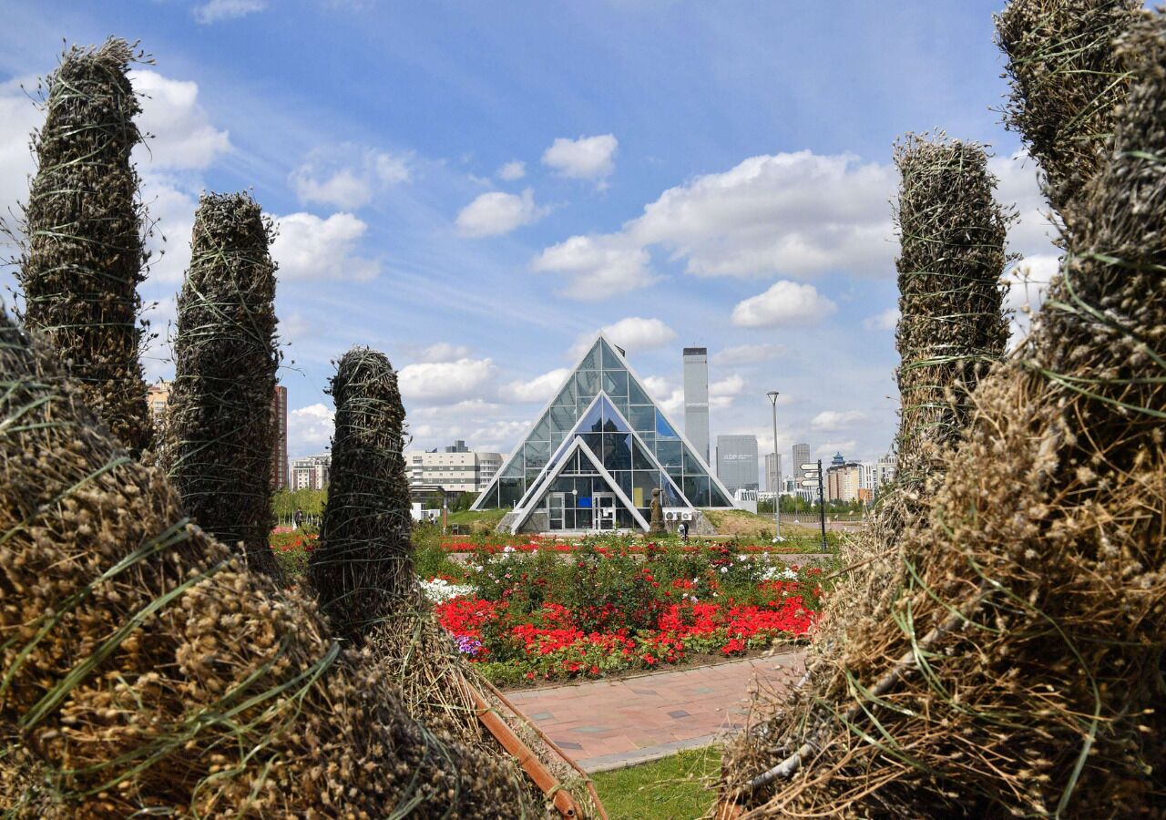A modern building with a triangular glass facade, surrounded by a landscaped garden with red flowers and tall, cylindrical structures.