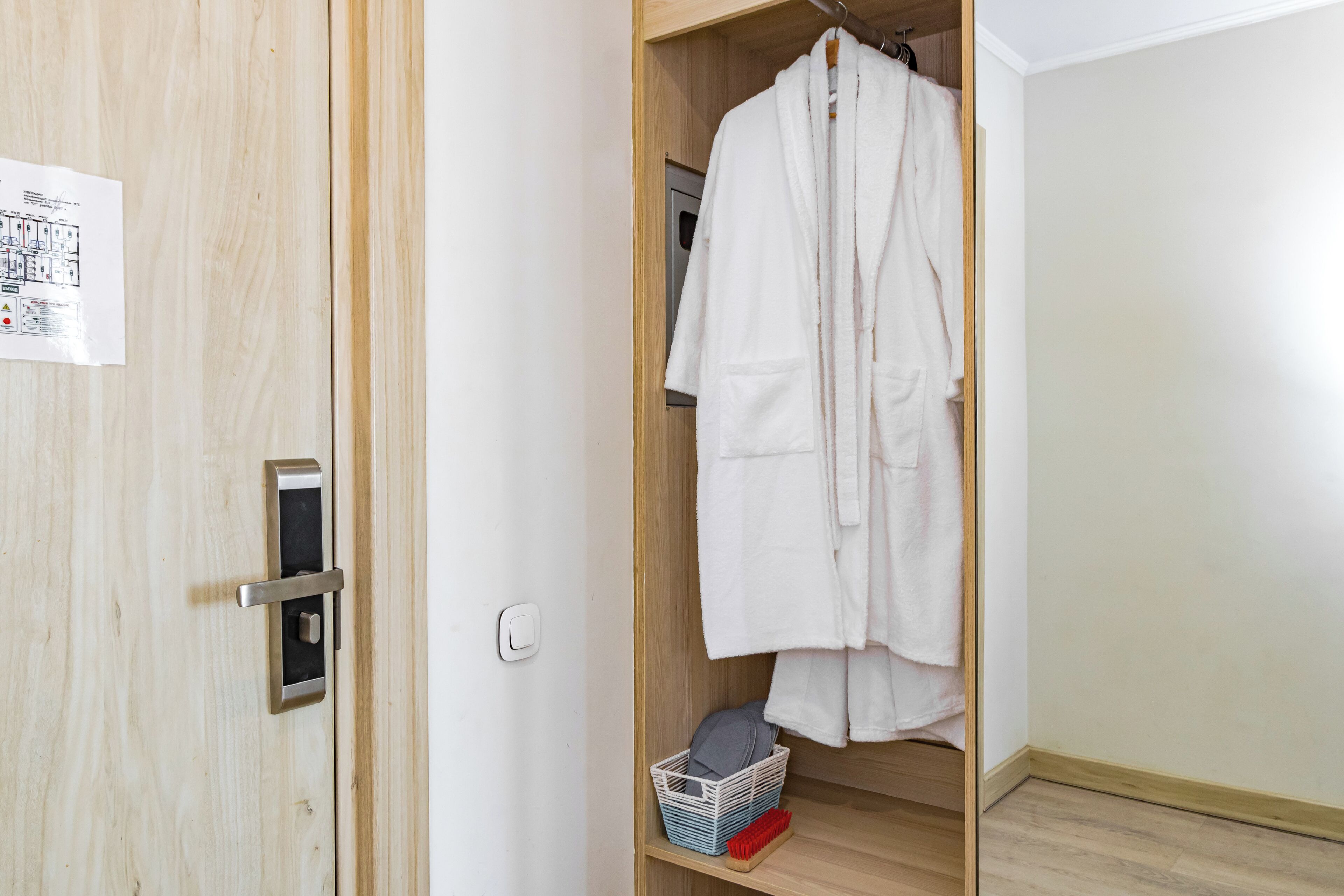 A wooden wardrobe with a white robe hanging inside, a small basket with a red brush, and a safety sign on the door.