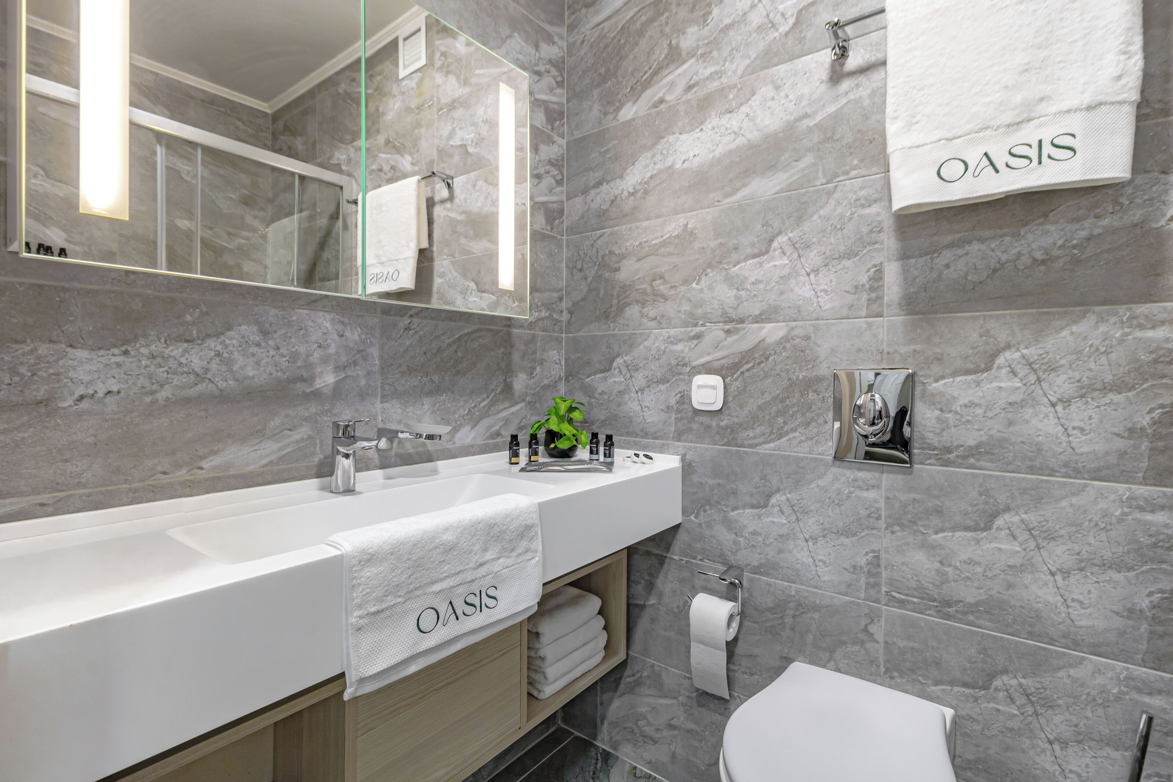A modern bathroom with a large mirror, a white sink with a towel labeled "OASIS," and a marble wall.