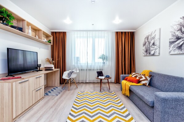 Una sala de estar moderna con un televisor de pantalla plana, estanterías de madera, un sofá gris, un tapete amarillo y gris, y un ventanal con cortinas.