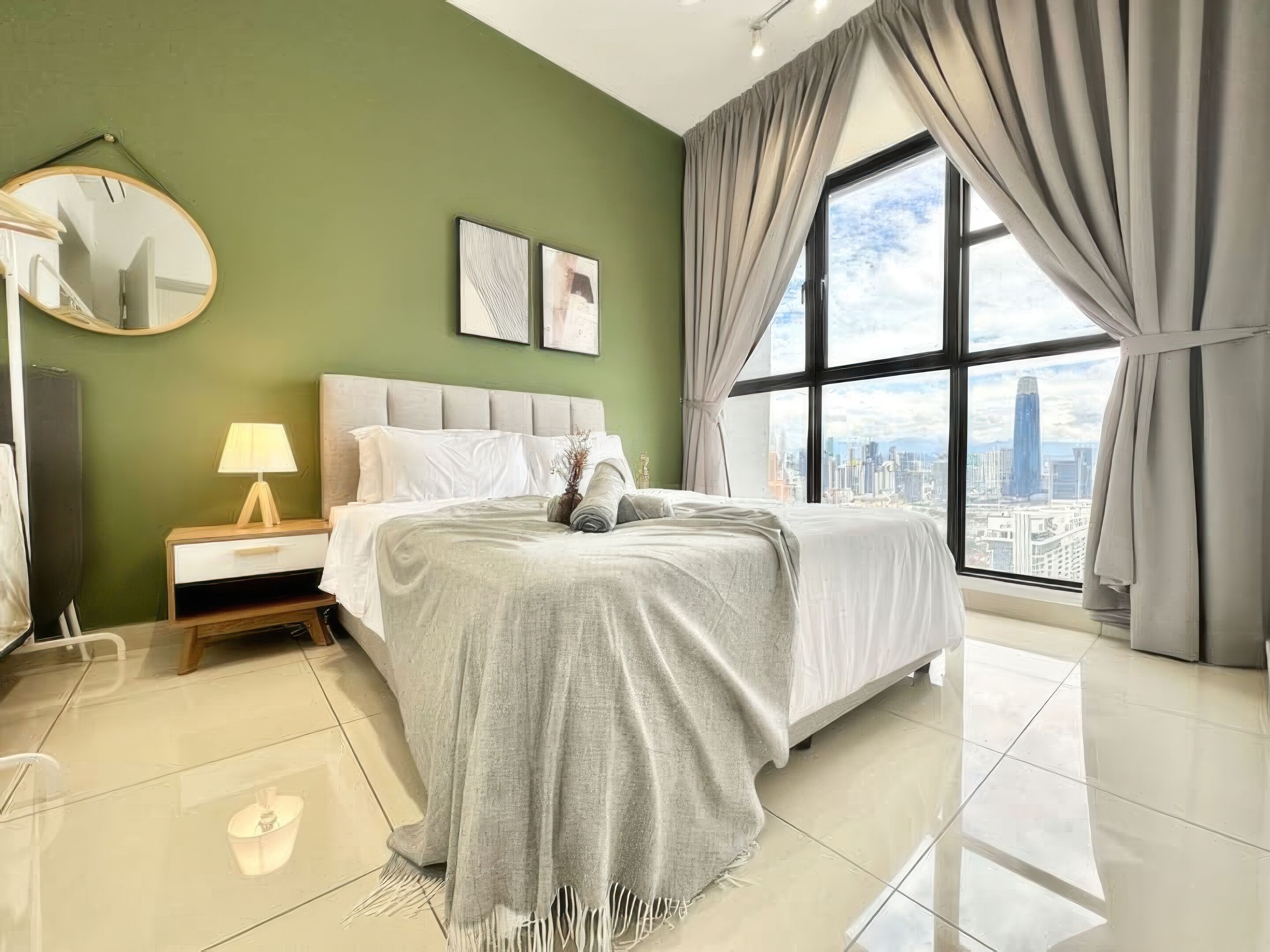 A modern bedroom with a large bed, a nightstand, a lamp, a mirror, and a city view through a large window.