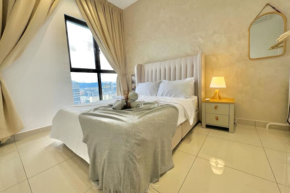 A bedroom with a bed, a nightstand, a lamp, a mirror, and a view of the city through a large window.
