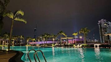 A nighttime swimming pool with palm trees and city buildings in the background.